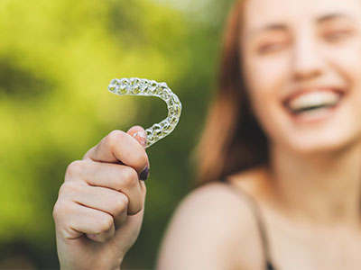 An image of a young woman holding up a transparent dental retainer with her left hand, smiling and laughing in the background.